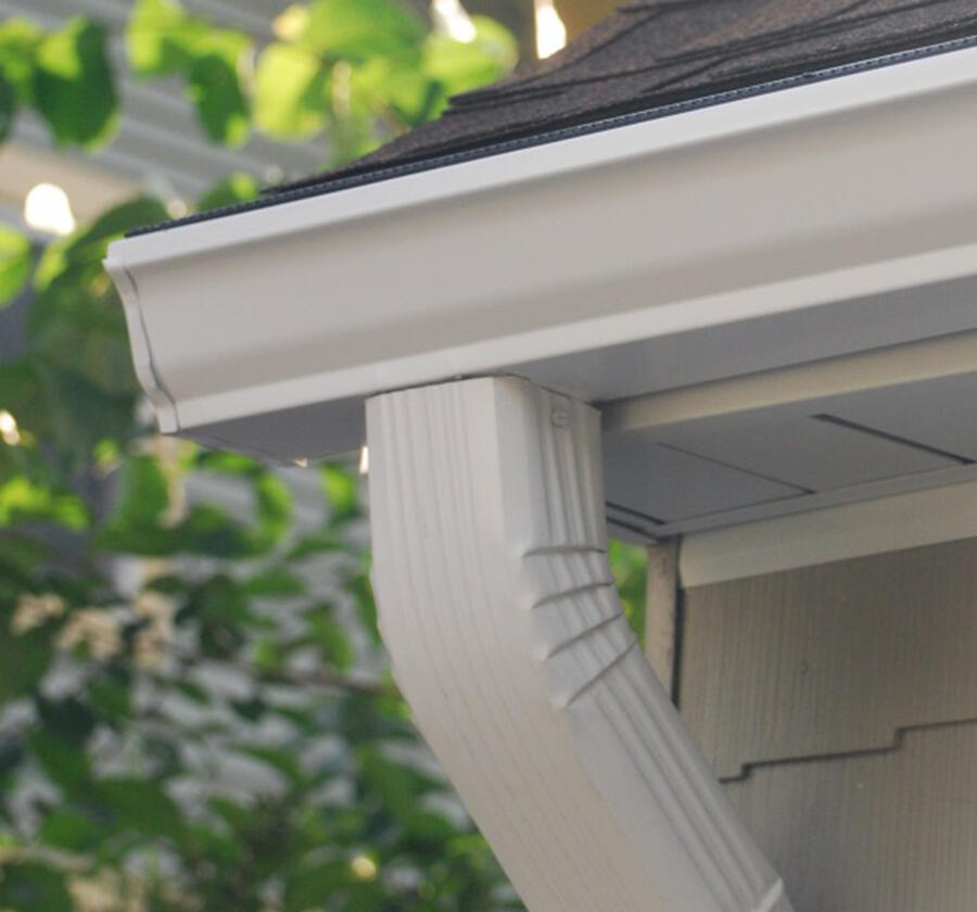 Close-up of a house's exterior corner showing white siding, gutter, and downspout, with green leaves in the background.